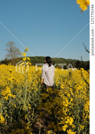 woman in white summer dress and straw hat walking through yellow rapeseed flower field under clear blue sky in countryside. High quality photo woman in white summer dress and straw hat walking through yellow rapeseed flower field under clear blue sky in countryside. High quality photo 125770346