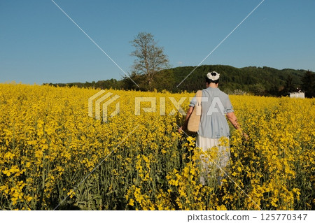 woman in white summer dress and straw hat walking through yellow rapeseed flower field under clear blue sky in countryside. High quality photo woman in white summer dress and straw hat walking through yellow rapeseed flower field under clear blue sky in countryside. High quality photo 125770347