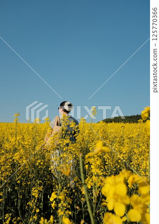 woman in white summer dress and straw hat walking through yellow rapeseed flower field under clear blue sky in countryside. High quality photo 125770366