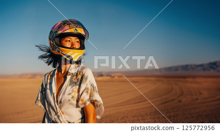 Beautiful young Japanese woman biker wearing helmet against sandy desert backdrop, independence defined by woman's fearless energy and daring desert adventures, copy space for active vacation concept Beautiful young Japanese woman biker wearing helmet against sandy desert backdrop, independence defined by woman's fearless energy and daring desert adventures, copy space for active vacation concept 125772956