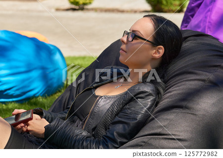 Young woman with glasses resting on street cushion pouf on a green lawn 125772982