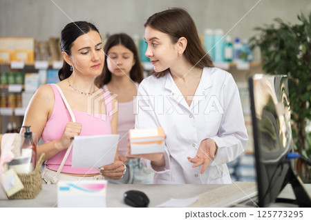 Young professional female parmacist working computer and helping mother and daughter clients with prescription in hand look for medicine in drugstore 125773295