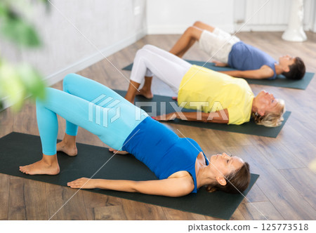 Middle-aged woman practicing bridge pose of yoga in light fitness room 125773518