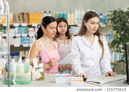 Young professional female parmacist working computer and helping mother and daughter clients with prescription in hand look for medicine in drugstore Young professional female parmacist working computer and helping mother and daughter clients with prescription in hand look for medicine in drugstore 125773613