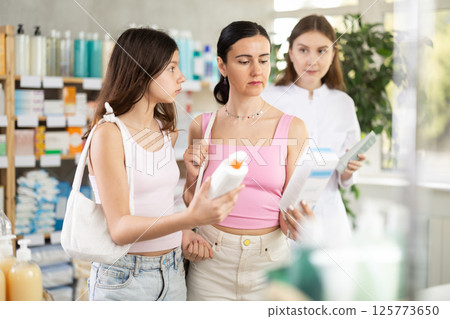 Focused mother and daughter choosing cream or skin care product in pharmacy, young woman pharmacist consulting and offering variety of goods to customer Focused mother and daughter choosing cream or skin care product in pharmacy, young woman pharmacist consulting and offering variety of goods to customer 125773650