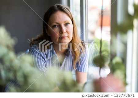Thoughtful brunette woman stands near casement, looks out window 125773671
