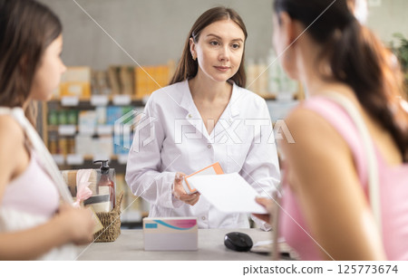 Portrait of woman pharmacist standing at prescription counter and helping mother and daughter choosing pill box and making decisions about pharmaceuticals and health care at pharmacy shop Portrait of woman pharmacist standing at prescription counter and helping mother and daughter choosing pill box and making decisions about pharmaceuticals and health care at pharmacy shop 125773674