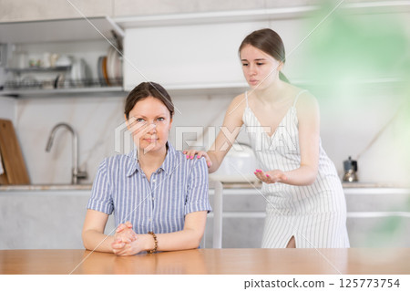Offended mother sitting at the kitchen table, daughter standing behind Offended mother sitting at the kitchen table, daughter standing behind 125773754