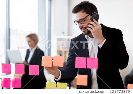 Young male professional wearing formal suit organizing tasks using sticky notes. Transparent glass board displays structured planning. Female colleague working with laptop in office environment. 125773879