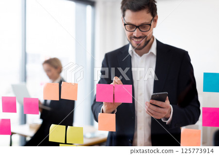 Adult male organizing tasks with colorful sticky notes on glass board, holding smartphone. Office backdrop with modern workplace vibes. 125773914