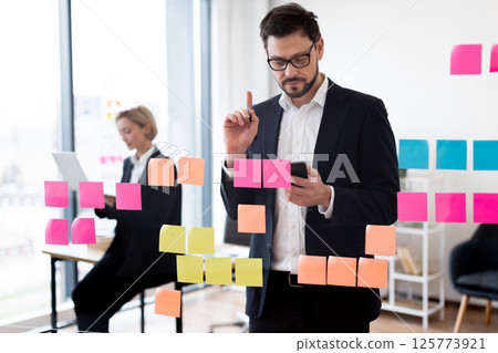 Business individuals, man and woman, collaborating on project using colorful notes on glass board. Scene depicts teamwork and planning in professional setting with modern office environment Business individuals, man and woman, collaborating on project using colorful notes on glass board. Scene depicts teamwork and planning in professional setting with modern office environment 125773921