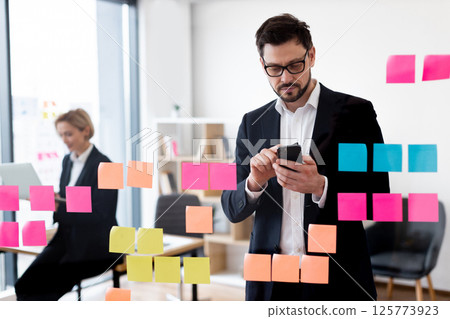 Young male and female professionals collaborating using post-it notes and gadgets for planning at office, representing teamwork, organization, and business strategy. 125773923