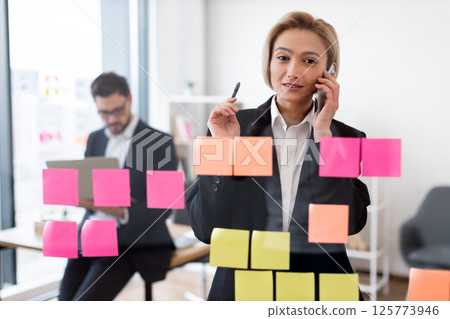 Confident adult businesswoman engaging in phone call, organizing work with colorful notes on transparent board in modern office. Male colleague visible working in background. Confident adult businesswoman engaging in phone call, organizing work with colorful notes on transparent board in modern office. Male colleague visible working in background. 125773946