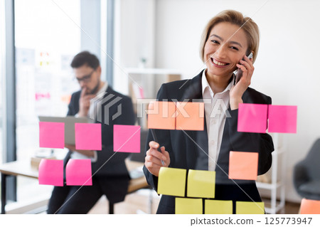 Businesswoman discussing strategy on phone near glass with sticky notes, colleague working background 125773947