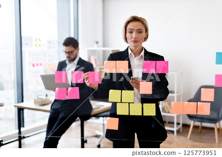 Businesswoman organizing tasks with colorful sticky notes on transparent glass board, office setting 125773951