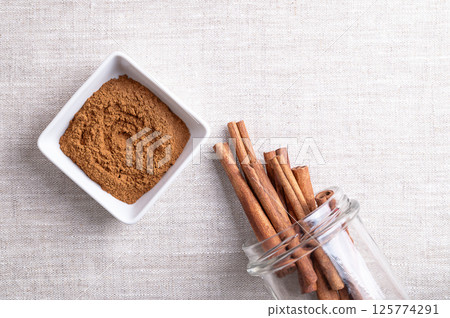 Cassia cinnamon powder in a white bowl, with cinnamon sticks sticking out of an open screw-top jar, on linen fabric, with empty space for text. Chinese cinnamon, Cinnamomum cassia, used as a spice. 125774291