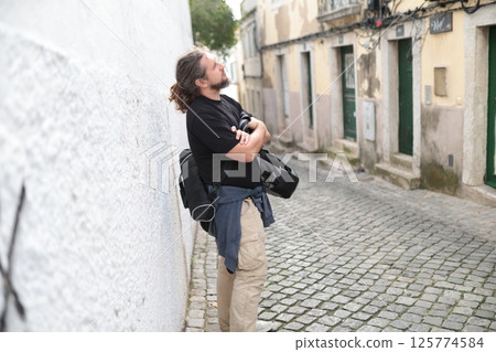 Photographer resting against white wall in cobblestone alleyway 125774584