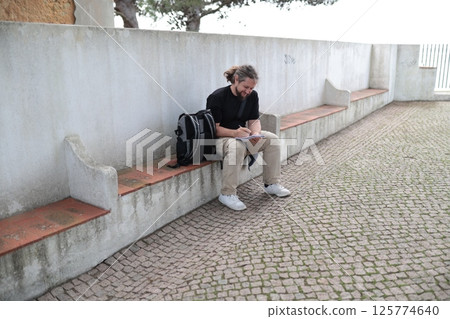 Smiling tourist writing notes on notepad while sitting on bench Smiling tourist writing notes on notepad while sitting on bench 125774640