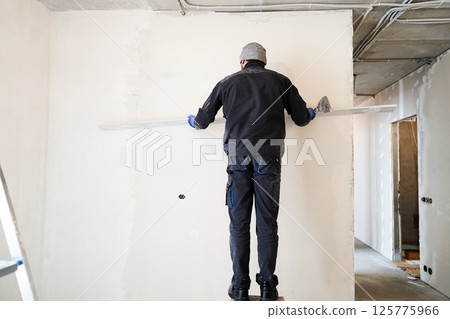 Applying gypsum plaster in a mechanized way to the wall. A worker removes excess plaster using a metal rule. High quality photo 125775966