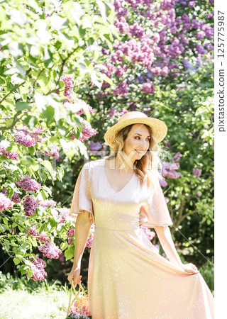 Young Woman In Summer Hat Enjoying The Fragrance Of Lilac Blooms On Sunny Day. lady in a straw hat and dress smelling lilac flowers in spring sunlit garden. Young Woman In Summer Hat Enjoying The Fragrance Of Lilac Blooms On Sunny Day. lady in a straw hat and dress smelling lilac flowers in spring sunlit garden. 125775987