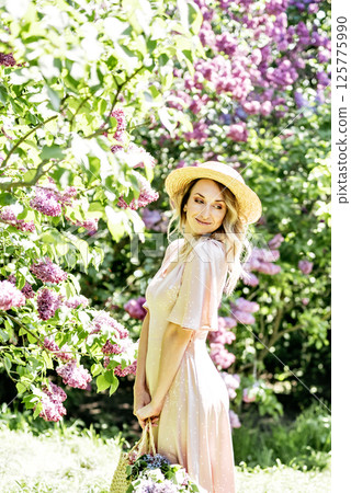 Young Woman In Summer Hat Enjoying The Fragrance Of Lilac Blooms On Sunny Day. lady in a straw hat and dress smelling lilac flowers in spring sunlit garden. Young Woman In Summer Hat Enjoying The Fragrance Of Lilac Blooms On Sunny Day. lady in a straw hat and dress smelling lilac flowers in spring sunlit garden. 125775990