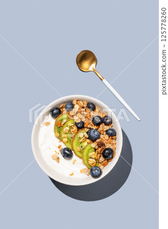 Natural yogurt with granola, kiwi and blueberries in a bowl on a blue background with spoon. 125778260