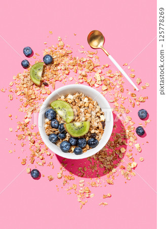 Baked granola or muesli in a bowl with kiwi and blueberries scattered on a pink background. Baked granola or muesli in a bowl with kiwi and blueberries scattered on a pink background. 125778269