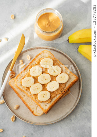 Peanut butter toast with banana on a plate on a light background with morning shadow. 125778282