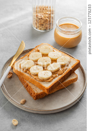 Peanut butter toast with banana on a plate on a light background with morning shadow. Peanut butter toast with banana on a plate on a light background with morning shadow. 125778283