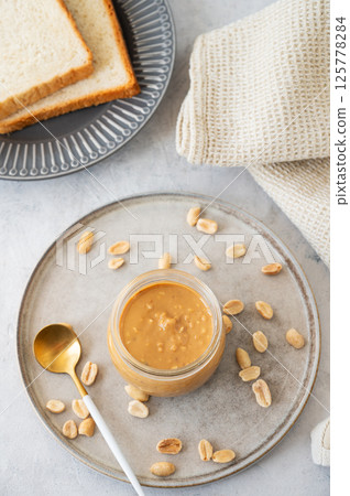 Peanut butter in glass jar on light background with salted nuts and bread toasts. Peanut butter in glass jar on light background with salted nuts and bread toasts. 125778284
