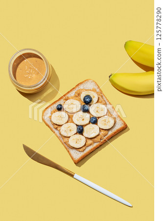 Peanut butter toast with banana, blueberry, nuts and knife on a yellow background. 125778290