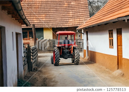 Farm tractor driving between houses on a village street Farm tractor driving between houses on a village street 125778513