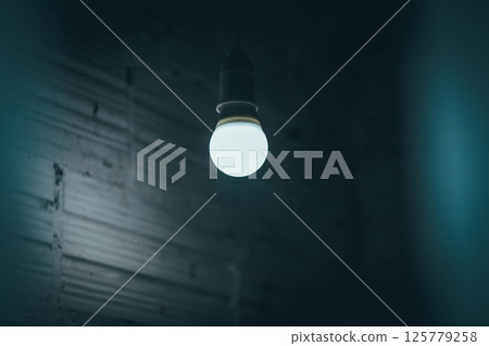 Single glowing LED bulb hanging from ceiling in a dark abandoned room 125779258
