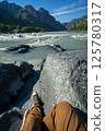 Man Sitting on Rocky Riverbank with Mountain View. Point Of View Shot. Adventure Travel Concept. 125780317