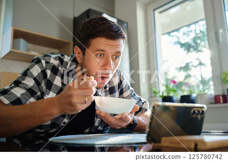 Surprised man has emotional reaction to phone while eating breakfast Surprised man has emotional reaction to phone while eating breakfast 125780742