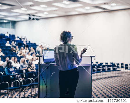 Female speaker giving a talk on corporate business conference. Unrecognizable people in audience at conference hall. Business and Entrepreneurship event 125780801