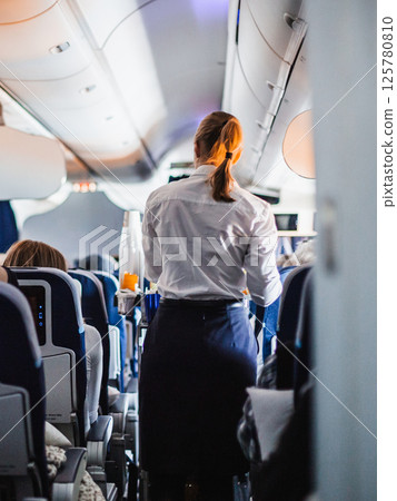 Interior of airplane with passengers on seats and stewardess in uniform walking the aisle, serving people. Commercial economy flight service concept Interior of airplane with passengers on seats and stewardess in uniform walking the aisle, serving people. Commercial economy flight service concept 125780810