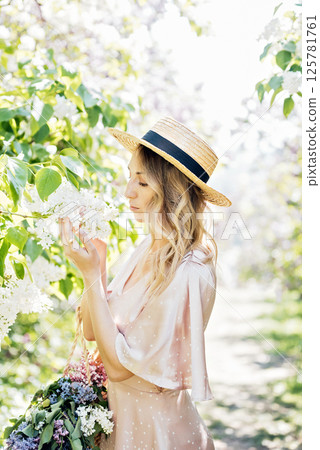 Woman in straw hat and pink dress gently smelling white lilac flowers in lush green garden on a sunny spring day Woman in straw hat and pink dress gently smelling white lilac flowers in lush green garden on a sunny spring day 125781761