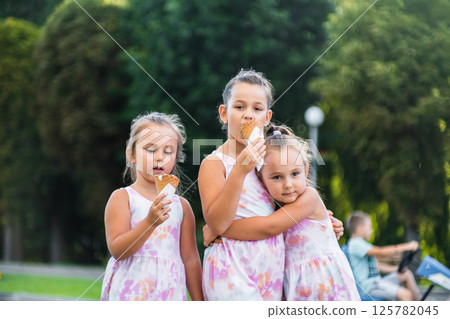 The sisters hold their melting ice cream cones, smiling at each other. The twins, matching in both dress and laughter, stand on either side of their older sister. 125782045