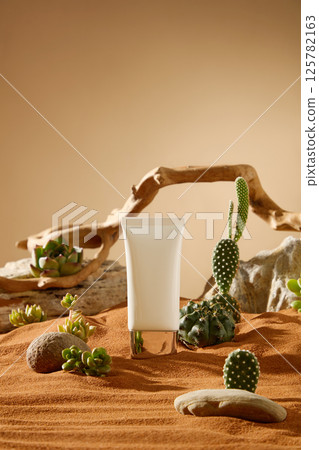 Mockup of white cosmetic tube with gold cap, standing on the center of the dry sand. Natural light shines in, shadows of objects fall on the sand. Spiny cactus grows around, interspersed with pebbles 125782163