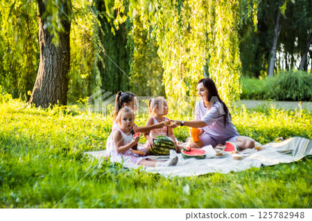 The girls and their mother enjoy a sunny picnic. The twins take big bites of watermelon, laughing. 125782948