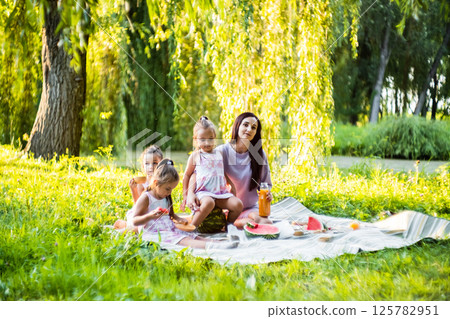 Sitting on the grass, the girls eat fresh watermelon with their mom. The twins joke, making their sister laugh. 125782951