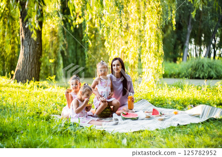 The three sisters sit around their mother, enjoying the picnic. The twins smile as they take bites of watermelon.  125782952