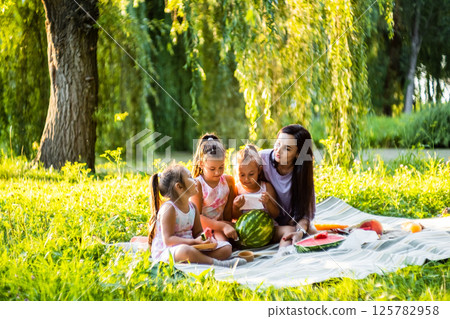 Under the warm sun, the family enjoys sweet watermelon. The twins make silly faces, making everyone laugh. 125782958