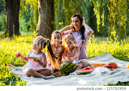 Under the warm sun, the family enjoys sweet watermelon. The twins make silly faces, making everyone laugh. 125782959