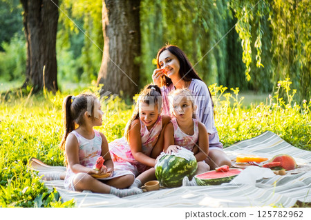 The mother watches her daughters enjoy the picnic. The twins hold big slices of watermelon, juice dripping. The mother watches her daughters enjoy the picnic. The twins hold big slices of watermelon, juice dripping. 125782962