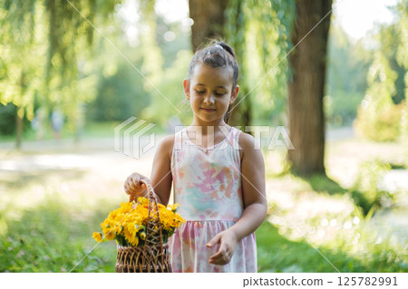 In a lovely park, a girl in a dress carries a basket of yellow flowers. 125782991