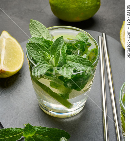 Glass of mojito cocktail with fresh mint leaves, lime slices, and ice cubes on a dark stone background. 125783109