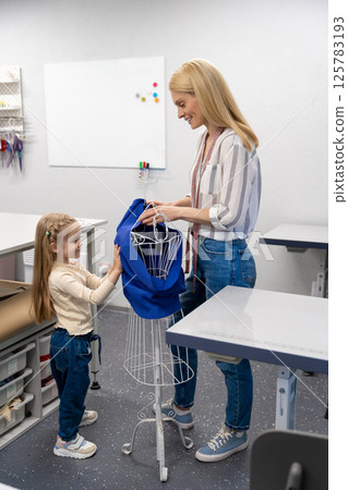 Blonde long-haired female dress designer in her studio with her daughter Blonde long-haired female dress designer in her studio with her daughter 125783193