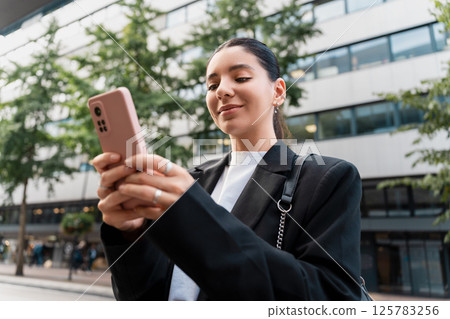 Confident woman scrolling through banking apps and map routes, blending finance and travel in a sleek street look Confident woman scrolling through banking apps and map routes, blending finance and travel in a sleek street look 125783256